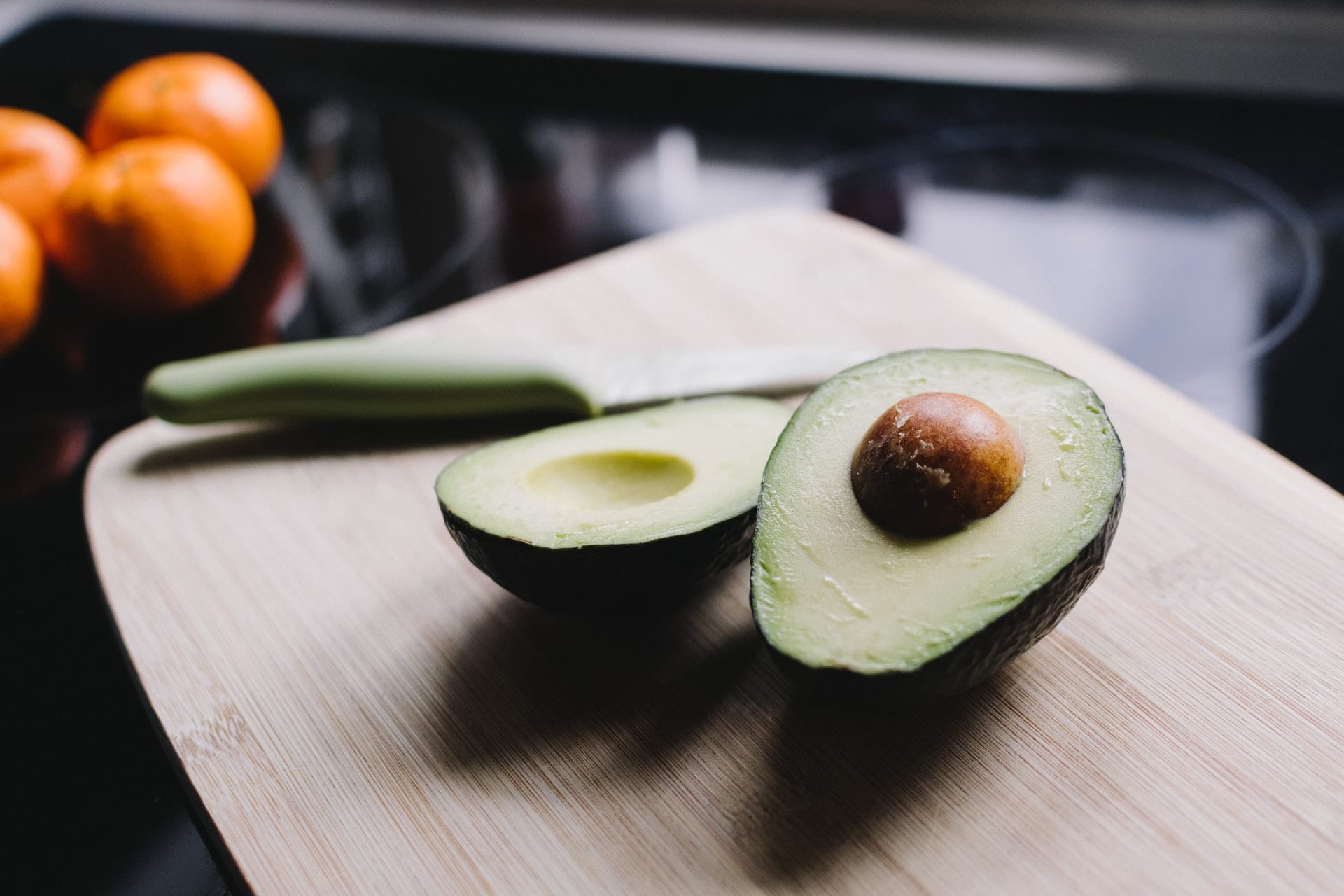 Avocado cut in half on a chopping board beside a knife, with oranges in the background